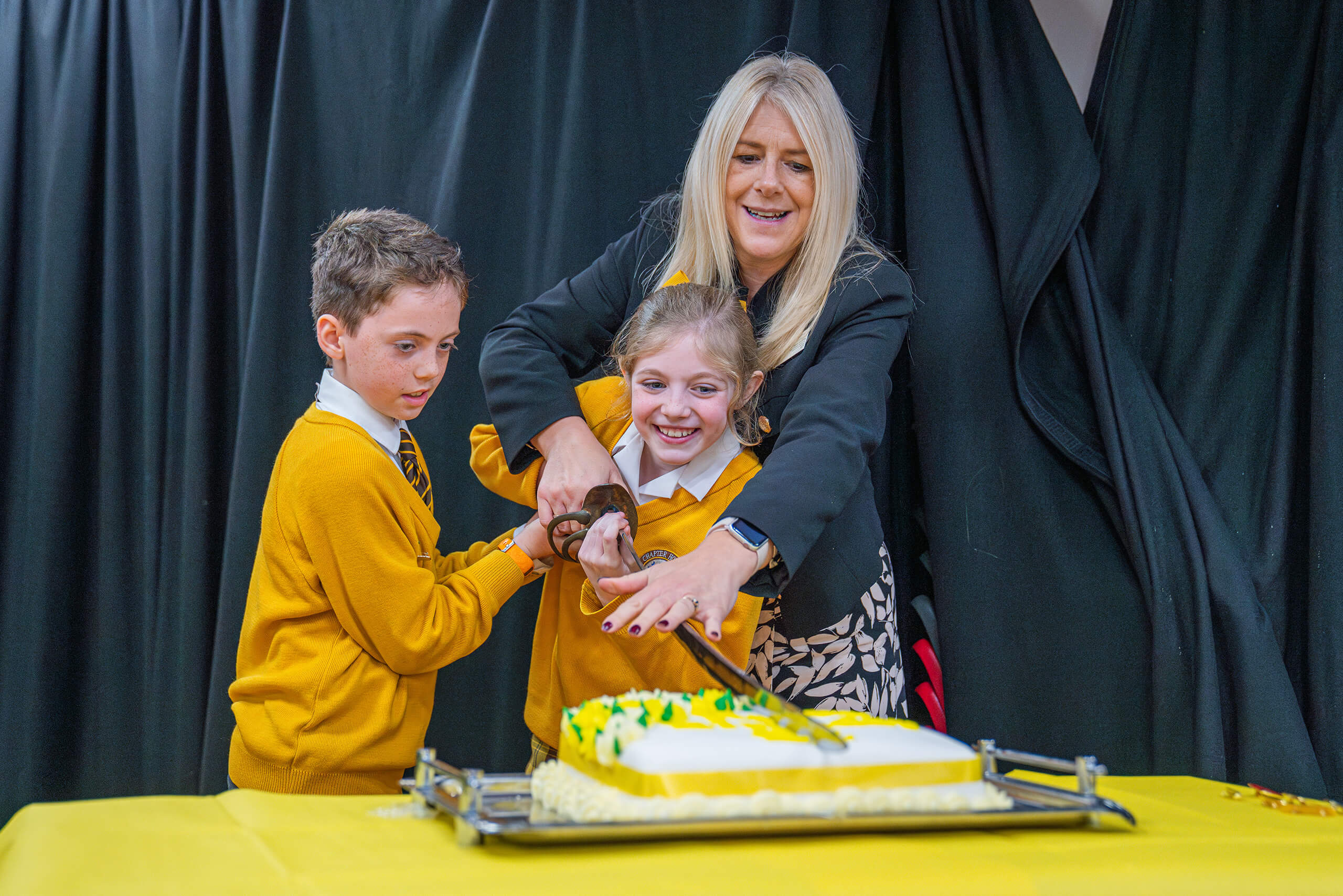 Two students and the Head of Chapter House cutting a birthday cake with a sword.